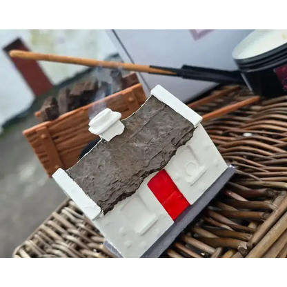 White cottage burner with a red door on a slate, smoke rising, turf cart behind on wicker